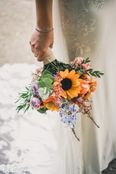 Strauß mit Sonnenblumen, bunten Blumen und grünem Blattwerk, in einer Hand gehalten.